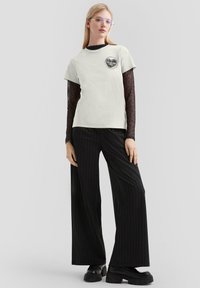 Helles graues T-Shirt mit rundem Grafiklogo, darüber eine dunkle, durchsichtige langärmelige Bluse. Weite schwarze Nadelstreifen-Hosen und klobige schwarze Stiefel.