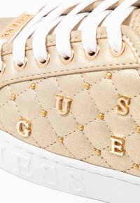 Sneaker oro trapuntato con lacci bianchi, occhielli dorati e borchie. Presenta la scritta "GUESS" in rilievo sul lato e una suola in gomma bianca.