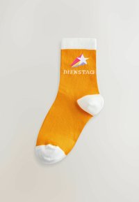Orange strømpe med hvidt tå, hæl og manchet, der har en hvid stjerne med pink og lilla spor og ordet "DIENSTAG" under.