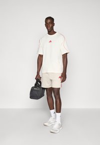 Sportowy strój składający się z kremowej koszulki z krótkim rękawem z czerwonymi akcentami i logo, beżowych spodenek, białych skarpet, szarych butów oraz czarnej torby sportowej.