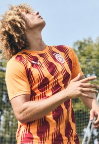 Maglia casalinga del Galatasaray con base arancione, strisce bordeaux e logo sul petto. Realizzata in tessuto traspirante, presenta maniche corte e colletto tondo.