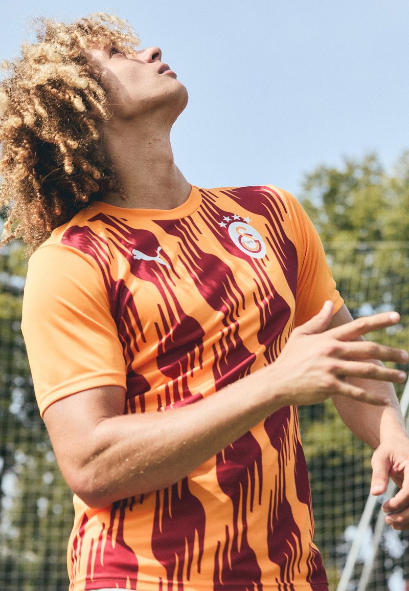 Maglia casalinga del Galatasaray con base arancione, strisce bordeaux e logo sul petto. Realizzata in tessuto traspirante, presenta maniche corte e colletto tondo.