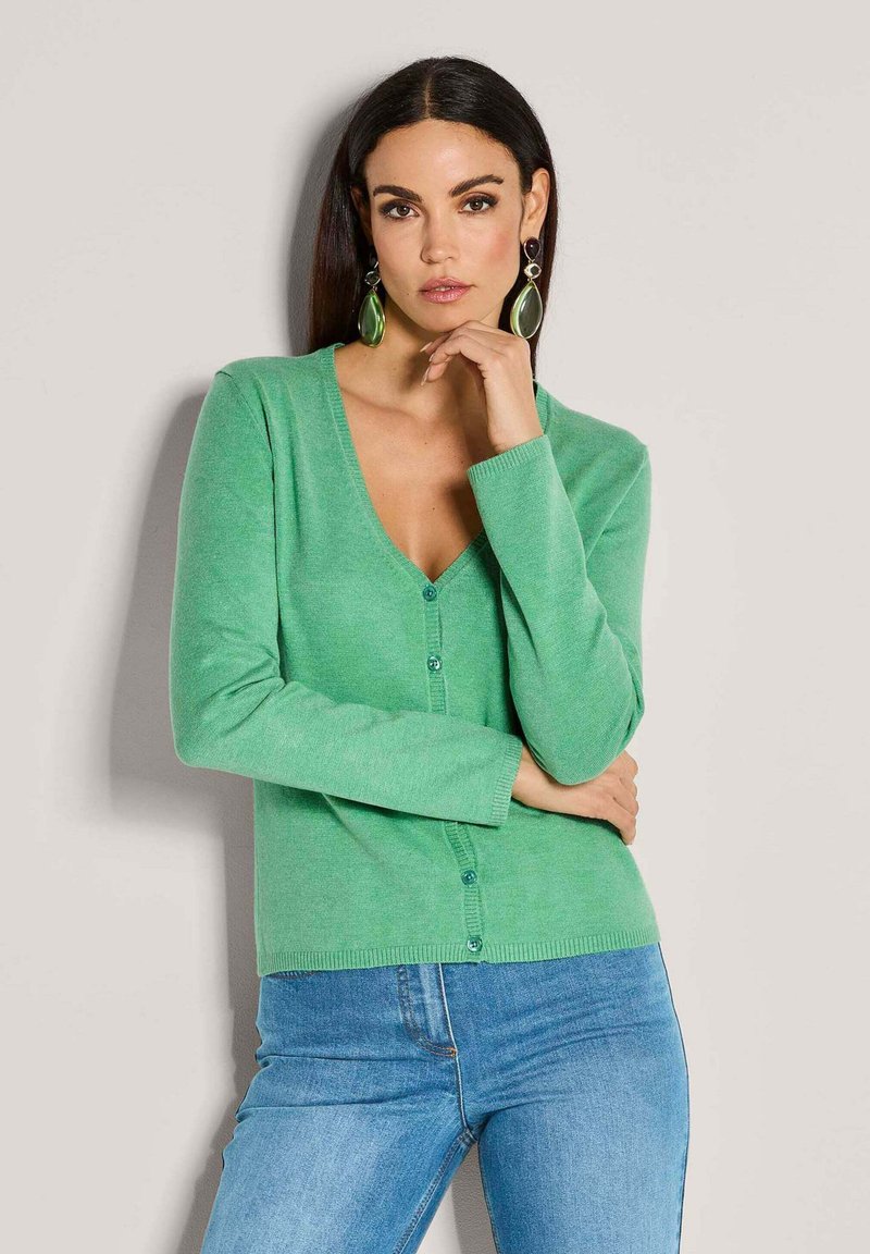 Groene v-hals cardigan van zacht materiaal, met een knoopsluiting en lange mouwen, gecombineerd met blauwe jeans.