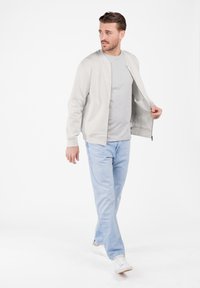 Hombre vestido con chaqueta gris clara sobre camiseta gris, jeans azul claro y zapatillas blancas, caminando mientras mira hacia un lado.