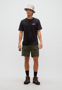 Άνδρας που φοράει μπεζ καπέλο τύπου bucket, μαύρο μπλουζάκι με γράφημα, ελαιδί cargo σορτς, μαύρες κάλτσες και καφέ παπούτσια ορειβασίας, κρατώντας γυαλιά ηλίου σε ένα χέρι.