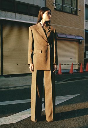 Donna in piedi sulla strada con un blazer marrone oversize e pantaloni a gamba larga, che guarda di lato vicino a persiane chiuse e coni stradali.
