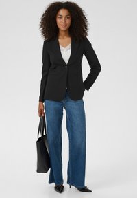 Blazer noir à un seul bouton, assorti à un haut en dentelle blanche et un jean bleu à jambes larges. Le mannequin tient un sac cabas noir et porte des escarpins pointus.