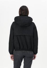 Μαύρο ελαφρύ μπουφάν με κουκούλα, με χαλαρή εφαρμογή, ελαστικούς καρπούς και ζωνάκι, εμφανιζόμενο από πίσω. Απλός σχεδιασμός, χωρίς μοτίβα.