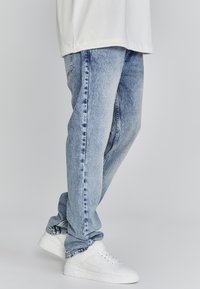 Jeans de denim azul claro con un corte relajado, con un lavado desgastado, costuras visibles y una textura ligeramente desgastada. Llevados con zapatillas blancas.