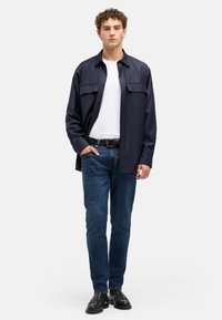 Marineblå skjorte med knapper og to brystlommer, båret over en hvid t-shirt, kombineret med mørkeblå jeans og sorte sko. Glat stoftekstur.