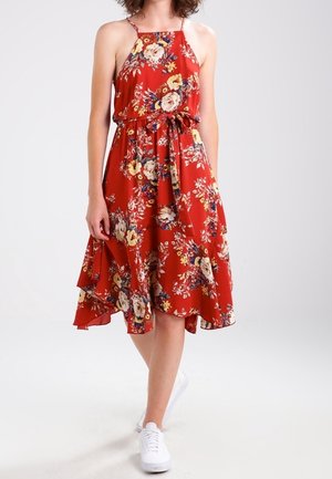 Kobieta w czerwonej sukience midi w kwiaty bez rękawów z paskiem w talii, w białych trampkach, stojąca na jednolitym tle.
