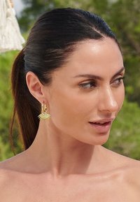 Orecchini a cerchio in oro con un design a raggi di sole e raggi testurizzati. Il modello ha lunghi capelli scuri, posizionato su uno sfondo verde sfocato.