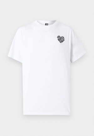 Joven con vitiligo que lleva una camiseta blanca de The North Face y pantalones marrones, de pie contra un fondo liso de color gris claro.