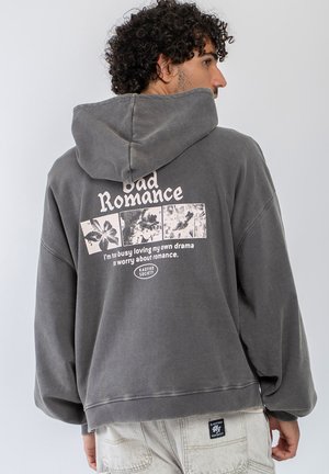 Mężczyzna z kręconymi włosami nosi ciemnoszarą bluzę z kapturem z napisem "Bad Romance" i kwiatowymi grafikami na plecach, w połączeniu z jasnymi dżinsami.