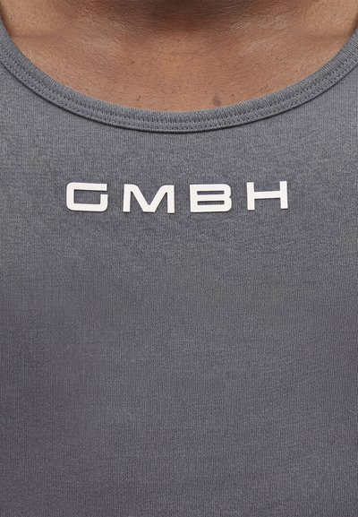 Κοντινό πλάνο ενός ατόμου που φοράει γκρι πουκάμισο με λευκά, έντονα γράμματα "GMBH" κεντραρισμένα κάτω από τη λαιμόκοψη.