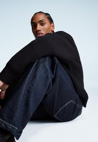 Czarny sweter z dzianiny o prążkowanej fakturze, w połączeniu z ciemnoniebieskimi dżinsami z kontrastowym przeszyciem i luźnym krojem. Zauważona pozycja siedząca.