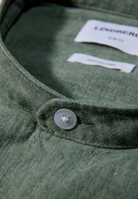 Cuello de camisa de lino verde con un borde cosido y un botón claro de cuatro agujeros. Las etiquetas son visibles en la zona del cuello.