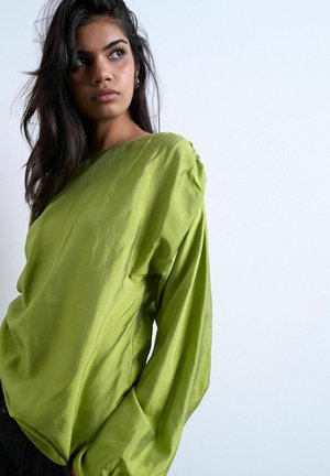 Jeune femme aux longs cheveux foncés portant un haut ample vert vif à manches longues, regardant vers la gauche sur un fond clair uni.