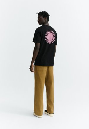 Homme debout, de profil, portant un t-shirt noir avec un motif circulaire rose dans le dos, un pantalon large couleur moutarde et des chaussures noires sans lacets.
