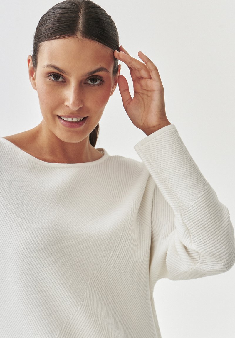 Biały sweter o textured z ukośnymi prążkami, szerokim dekoltem i opadającymi ramionami. Modelka lekko dotyka czoła.