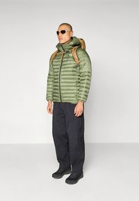 Grön vadderad jacka med dragkedja fram, grå byxor och svarta stövlar. Accessoarer inkluderar solglasögon och en brun ryggsäck med remmar.