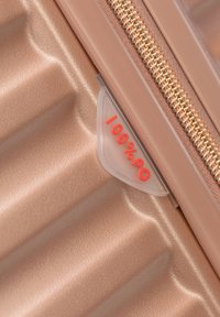 Esquina de maleta de bronce con textura brillante, con cremallera dorada y etiqueta translúcida que muestra "100% pc" en texto rojo.