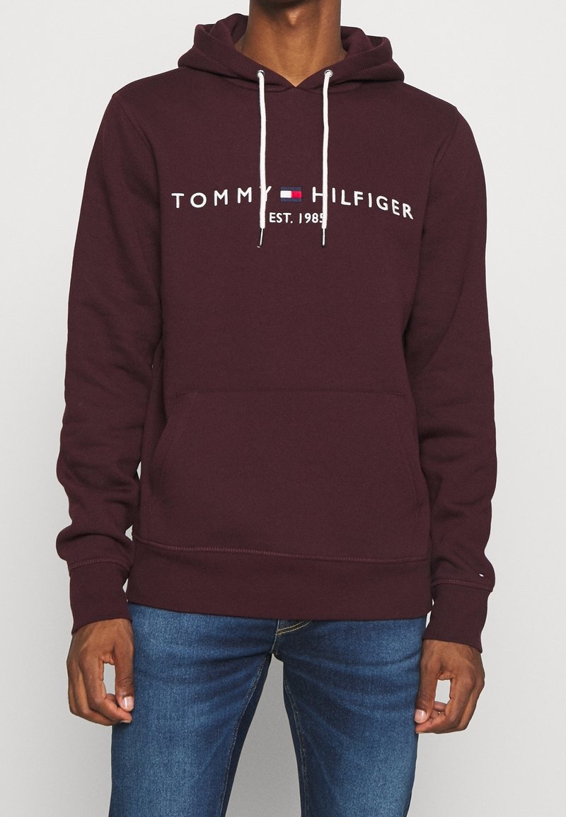 Homme portant un sweat à capuche Tommy Hilfiger bordeaux avec des cordons blancs et un logo sur la poitrine, associé à un jean bleu.