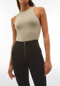 Body beige senza maniche abbinato a leggings neri a vita alta con cerniera frontale; texture liscia e design aderente.