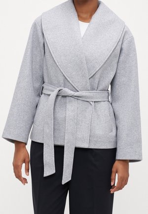 Kobieta ubrana w jasnoszary płaszcz zapinany na zakładkę z szerokimi klapami, długimi rękawami i przewiązanym paskiem, nosząca czarne spodnie.