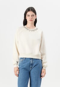 Felpa color crema con scollatura decorativa in perline, polsini e orlo a coste, caratterizzata da una vestibilità morbida e maniche raglan.