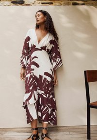 Maxikleid mit tiefem V-Ausschnitt, weißem Stoff mit großen kastanienbraunen Blättermustern und gestuften Rüschensaum. Schwarze Sandalen mit Riemchen runden das Outfit ab.
