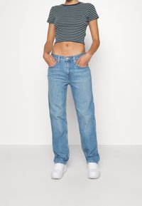 Randig kortärmad t-shirt i marinblått och grönt, kombinerad med ljusblå raka jeans. Modellen har vita sneakers på sig och händerna i fickorna.