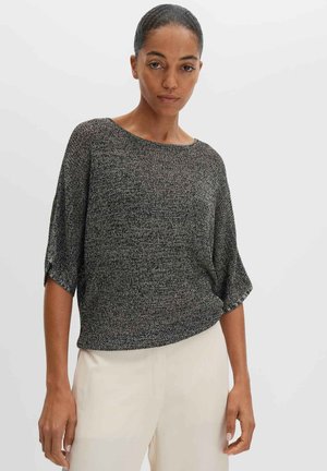 Femme aux cheveux foncés en chignon portant un haut en maille noir et blanc à manches courtes et amples et un pantalon beige clair sur un fond uni.