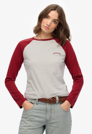 Mujer con cabello castaño hasta los hombros, vistiendo una camiseta de manga larga gris y roja con el texto "Superdry", pantalones a rayas y un cinturón trenzado marrón.