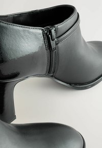 Cizme negre până la gleznă cu un finisaj lucios, având un fermoar pe lateral și un toc moderat. Materialul pare să fie neted, cu variații subtile de textură.