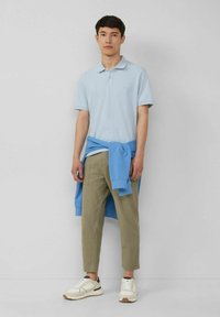 Helles blaues Poloshirt, olivgrüne schmal zulaufende Hose, blauer Pullover um die taille gebunden und weiße Turnschuhe mit schwarzen Akzenten.