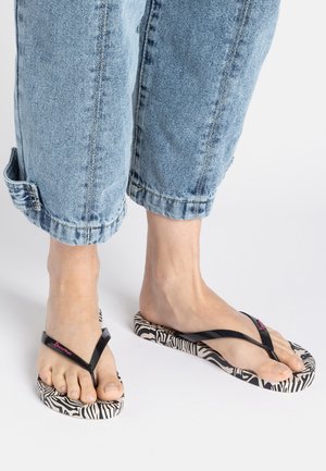 Schwarze Flip-Flops mit einer Zebramuster-Sohle, die einen glanzvollen Riemen und pinkes Logodesign aufweisen, getragen zu knöchellangen hellblauen Denim-Hosen.