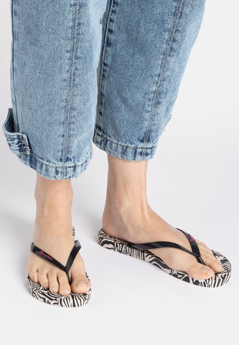 Black flip-flops with a zebra-print sole feature a glossy strap and pink logo detail, worn with cropped light blue denim pants.
