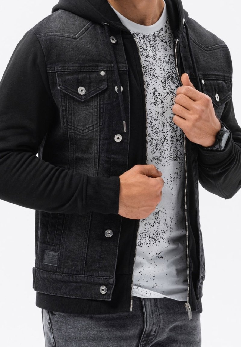 Homme portant une veste en jean noire avec capuche, un t-shirt blanc moucheté, un jean gris et une montre noire au poignet, réglant la fermeture éclair de la veste.