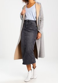 Abrigo largo gris con detalles beige, blusa azul claro, falda vaquera oscura de cintura alta y botas blancas hasta el tobillo. La falda tiene una silueta ajustada.