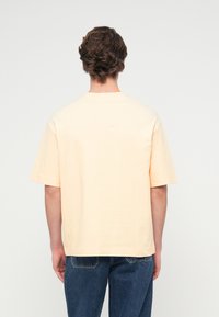 Κοντομάνικο, ανοιχτό ροδακινί T-shirt κατασκευασμένο από βαμβάκι. Σχέδιο με στρογγυλή λαιμόκοψη και άνετη εφαρμογή, με απλή πλάτη και χωρίς επιπλέον λεπτομέρειες.