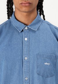 Denim button-up shirt in light blue with a collar, silver buttons, and a left chest pocket featuring white embroidered "obey" text.
