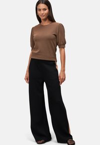 Haut court texturé marron à manches courtes avec épaules bouffantes, associé à un pantalon noir à jambes larges avec fentes latérales et des chaussures à motifs.