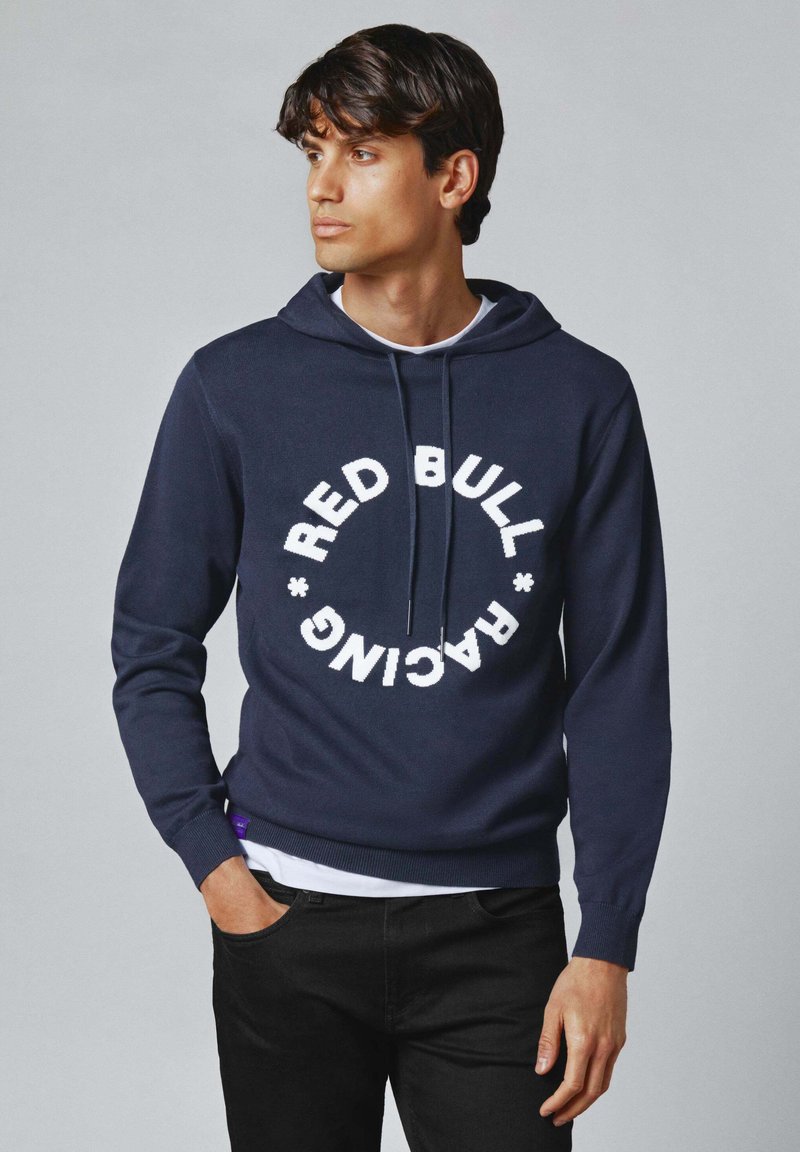 Tumši zils kapuci džemperis, izgatavots no mīkstas auduma, ar apaļu baltu "RED BULL RACING" logo. Tam ir priekšējā kabata un regulējams auklas kapucis.