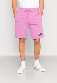 Pantalones cortos de algodón rosa con bolsillos laterales, cinturilla elástica y cordón. Presenta un logo azul de "GAP" en la pierna izquierda.