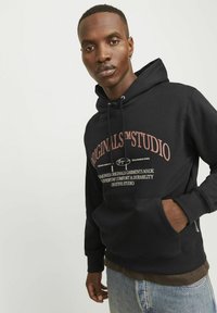 Sudadera negra con bolsillo frontal, capucha con cordón y texto impreso en colores claros. Presenta un corte relajado y una tela texturada.