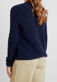 Mörkblå stickad tröja, strukturerat ullmaterial, ribbad halsringning, avslappnad passform, sidofickor, kombinerad med ljusbeiga byxor.