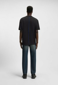 T-shirt nera a maniche corte, con una cucitura verticale blu lungo il centro della schiena, abbinata a pantaloni in denim scuro larghi e scarpe nere.