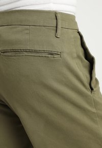 Ingrandimento di pantaloni verde oliva che mostrano le tasche posteriori e laterali, il passante della cintura e parte di una camicia bianca infilata dentro.