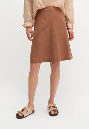 Frau trägt einen knielangen braunen Rock, ein beiges Langarmshirt und bequeme beigen Sandalen mit Schnallen, steht vor einem einfarbigen Hintergrund.